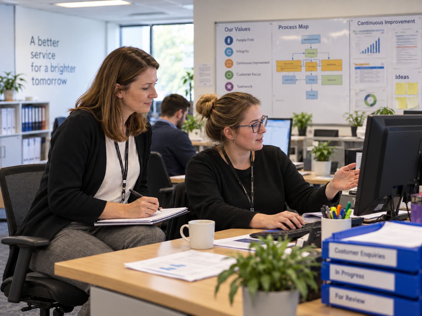 Twee collega’s aan een bureau bekijken samen een scherm, achter hen een procesoverzicht aan de wand